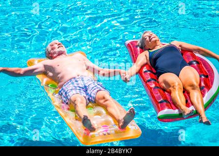 Old people senior couple relax and sleep on the blue swimming pool clear water lay down on trendy coloured inflatabler mattress lilos and taking hands with love for forever together lifestyle concept Stock Photo