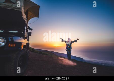 Travel and happiness concept for wanderlust people - woman with coloured warm clothes enjoy freedom and sunset near a car with tent on the roof - independence and wild traveler lifestyle female Stock Photo