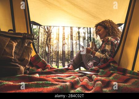 Cheerful happy people caucasian woman use cellular phone sitting outside a tent in the forest with sunlight in background - alternative travel vacation concept for independent adult with technology Stock Photo