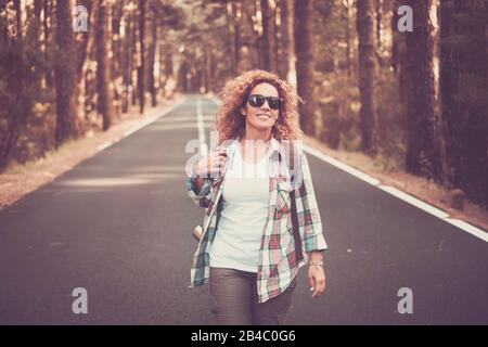 Cheerful happy free woman traveler people walking in the middle of a long road with forest and trees around - scenic travel concept - beautiful caucasian curly female enjoying the outdoor leisure activity Stock Photo