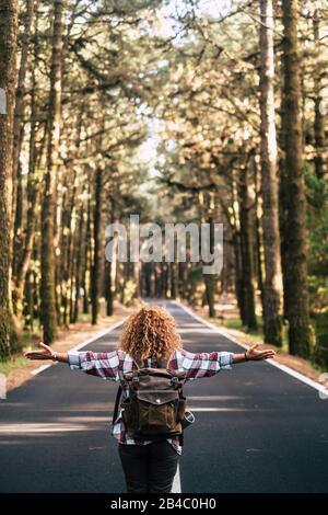 Alternative travel concept with curly hipster woman viewed from back open his arms and feel the freedom of the outdoors nature standing in the middle of a ling road - happiness and wanderlust lifestyle Stock Photo