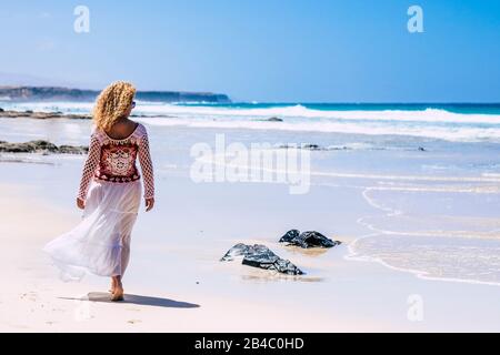 fashion trendy blonde curly woman walking on the white sand tropical resort beach with blue ocean in background - travel and summer holiday concept for beautiful people enjoying the outdoor Stock Photo