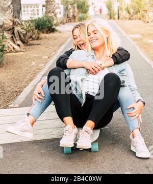Happy young people couple of friends girls have fun together riding a skateboard on the road for outdoor leisure funny activity - friendship and craziness lifestyle concept for alternative millennial Stock Photo