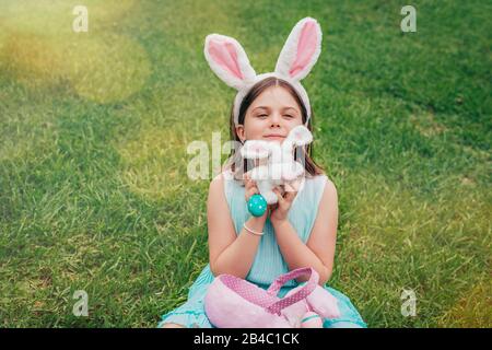 Cute little girl is wearing bunny ears on Easter day playing with colored eggs and bunny toy sitting on grass in garden. Stock Photo