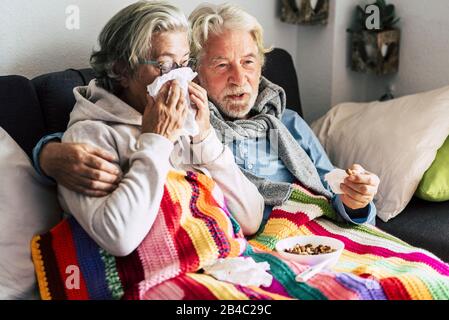 Couple of old aged senior people at home with seasonal winter cold illness disease  sit down on the sof together forever - health problems for retired man and woman with white hair Stock Photo