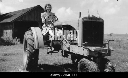 A woman driving a farm tractor, in front of a shed. This is a  c.1933 Allis-Chalmers farm tractor, with a hand crank up front and a belt-driver on the right side. (These tractors were built in West Allis, Wisconsin, near Milwaukee.) Two small American flags are on the front end. The young lady is wearing a 1940s-type casual day dress. This unknown woman is more posing than working, I think. Location is unknown.  To see my other vintage images, Search:  Prestor  vintage  woman  vehicle Stock Photo