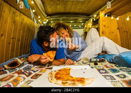 Romantic people adult young couple in love enjoy the little tiny house inside an old restored vintage van eating pizza together and having fun - concept of travel and alternative vacation Stock Photo