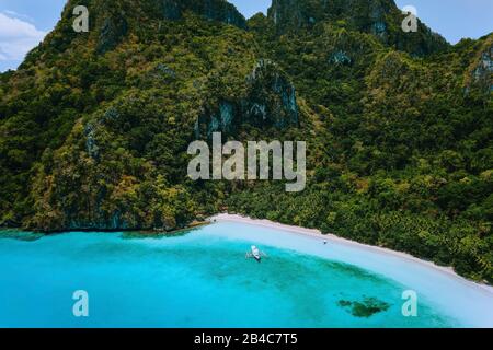 Aerial drone view of uninhabited tropical island with rugged mountains, rainforest jungle, sandy beach and tourist banca boat in blue lagoon. Stock Photo