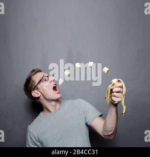 male person with glasses and heather gray t-shirt  eating banana. pieces of levitating fruit floating into mouth in front of gray studio background Stock Photo