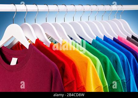 row of many fresh new fabric cotton t-shirts in colorful rainbow colors hangng on clothes rail in wrdrobe. Various colored shirts on blue white backgr Stock Photo