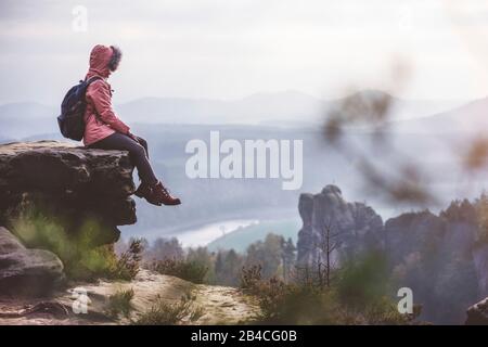 Junge Frau in winterlicher Kleidung mit Rucksack, sitzt am Rand der Klippe und genießt die Aussicht über Berge, Tal und Fluss, Reise-Lifestyle-Konzept Stock Photo