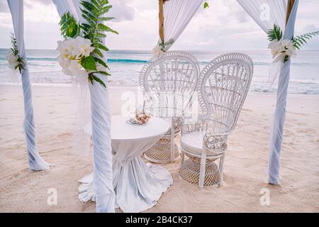 Decorated romantic wedding setting with table and chairs on sandy tropical beach with ocean and cloudy sky, Seychelles islands. Stock Photo