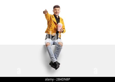 Trendy young man holding a box of popcorn, sitting on a panel and pointing isolated on white background Stock Photo