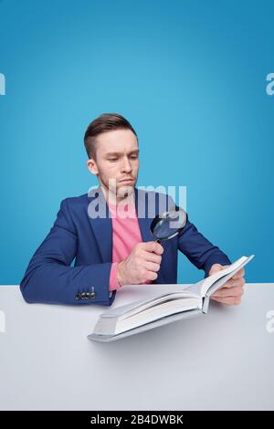 Serious male reader with magnifying glass sitting by desk and looking through text on page of book on blue background Stock Photo