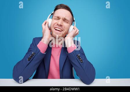Cheerful and excited young man with headphones enjoying his favrite music while sitting by desk on blue background Stock Photo