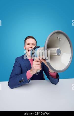 Angry young male manager or boss shouting in megaphone while making announcement or warning against blue background Stock Photo