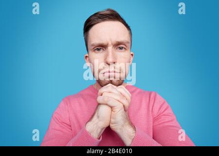 Upset young man in pink pullover keeping his hands by chin while thinking of what to do in front of camera over blue background Stock Photo