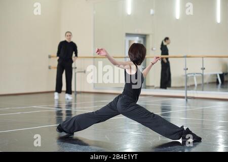 Young fit and flexible woman in black activewear and sneakers doing stretching exercise while training in dance studio Stock Photo