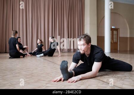 Young flexible man in black activewear sitting in twine position while training on the floor with group of dancers on background Stock Photo