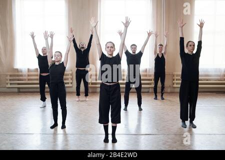 Group of young dancers in black activewear raising arms while standing on the floor against window in modern dance studio Stock Photo