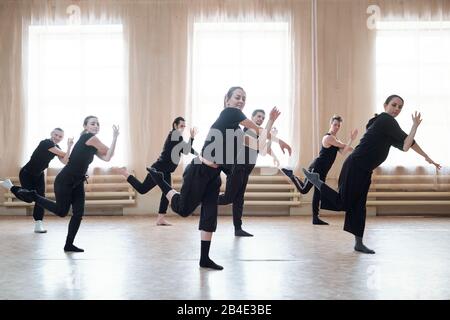 Seven young active men and women in black sportswear standing on the floor while doing dance exercise in large modern studio Stock Photo