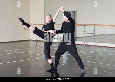 Young woman stretching one leg forward while holding by hands with her trainer during dance exercise in large studio Stock Photo