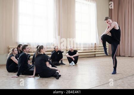 Group of several young dance course students sitting on the floor while looking at guy in activewear doing exercise Stock Photo
