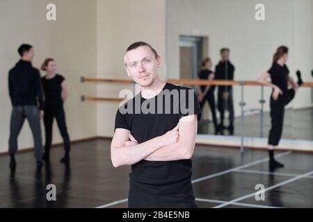 Young serious dancing instructor crossing arms by chest while standing in front of camera with students on background Stock Photo