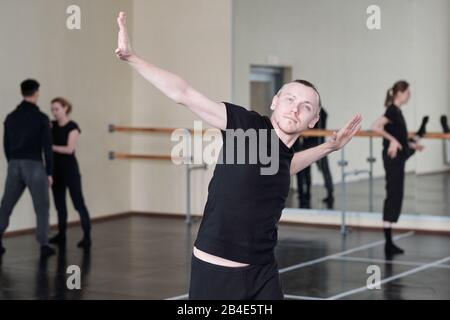 Young fit blond man in black activewear doing one of dance exercises while training in front of camera in studio of modern dancing Stock Photo