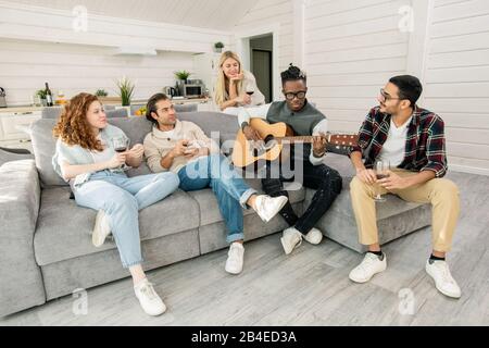 Young Africam man playing guitar while sitting on sofa among his friends listening to music while relaxing in living-room Stock Photo