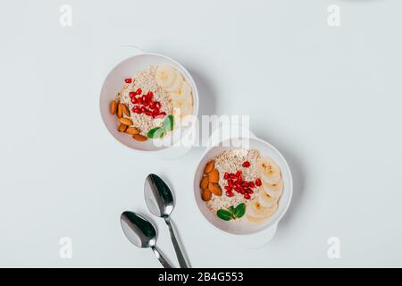 Served healthy breakfast for two of oatmeal with almonds, pomegranate seeds and banana slices, top view on white table. Stock Photo