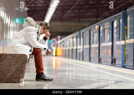 Crying man in white parka very upset, holding smartphone, gets bad news, holds forehead with his hand, sitting on bench in subway station. Problem in Stock Photo