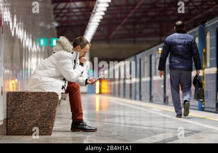 Crying young man in depression looking at phone gets bad news, receiving message/sms, covering mouth with hand, sitting on bench in subway station, pa Stock Photo