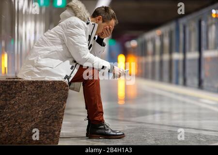 Crying man in white parka very upset, holding smartphone, gets bad news, covers his face with his hand, sitting on bench in subway station. Problem in