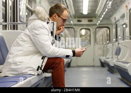 Crying young man looking at phone gets bad news, receiving message/sms, covering mouth with hand, sitting on seat in empty subway train, railway carri Stock Photo