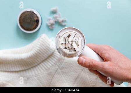 Man's hand holding a lint remover from a white acrylic or wool sweater. Electric device after cleaning and collected fluff/lint, light wooden backgrou Stock Photo