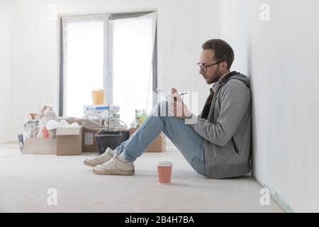 Man having a coffee break from renovating, sitting on the floor and writes upcoming purchases in his notebook, resting after work, soft focus. Home re Stock Photo