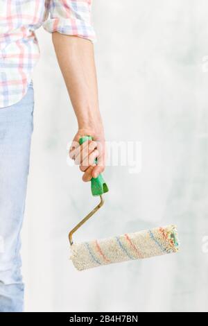Cropped image of man/painter in casual clothes holding a dirty roller for painting walls indoors. Closeup of hand with paint roller at home, vertical. Stock Photo