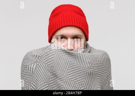 Sick man in red beanie wrapped in warm beige knitted plaid, suffering from cold, looking at camera, isolated on gray background. Eyes are red and sore Stock Photo