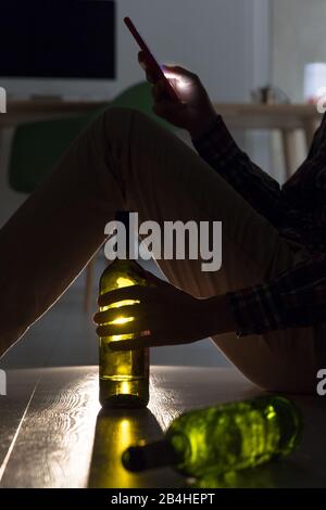 Closeup of alcoholic man sitting on the floor with a bottle in his hand, broke up with his girlfriend, typing message on smartphone at night. Alcohol Stock Photo