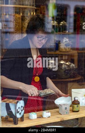 Blick durch eine Schaufensterscheibe auf ein Frau. Sie füllt Getreide in eine weiße Schüssel, Detail aus einem Unverpacktladen. Stock Photo