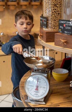 Eine Junge steht in einem Unverpacktladen an einem Regal mit Abfüllbehältern für Cerealien. Er wiegt Haselnüsse ab. Stock Photo