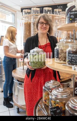 Eine Frau hält in einem Unverpacktladen ein Glas mit Süßigkeiten in den Händen. Im Hintergrund steht eine junge Kundin an einem Regal mit Abfüllbehält Stock Photo