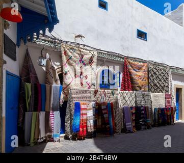 Colourful fabrics,carpets and homespuns hanging on the typical white and blue house wall for sale in the street market of Medina.Essaouira,Morocco. Stock Photo