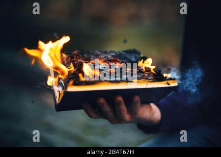 The sorcerer's hand holds a thick hardcover magic book that burns with a bright fire in the middle of the night and emits sparks and ashes into the ai Stock Photo