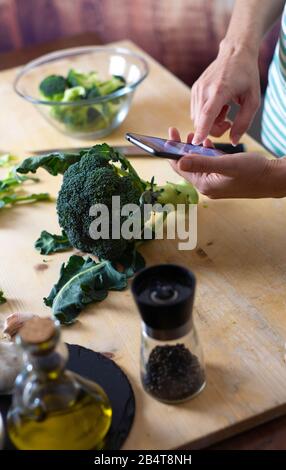 Moments of daily life in the Mediterranean cookery: young female cook consult her smartphone to find a recipe for broccoli on a wooden chopping board Stock Photo
