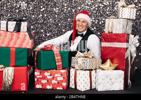 Smiling man in suit wearing Santa Claus hat posing with Christmas gift surrounded by snow Stock Photo