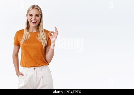 Confident girl got everything under control. Unbothered good-looking modern blond woman in orange t-shirt, with tattoos, show okay confirmation Stock Photo