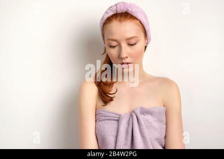 A red-haired girl in a lilac towel with a rim on her hair stands on a white background and looks down. Stock Photo