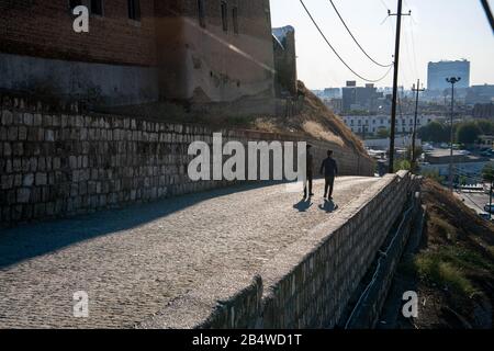 Iraq, Iraqi Kurdistan, Arbil, Erbil. Two men are walking donw a road. On the left is the Qalat citadel. In the background a view of Erbil at sunset. Stock Photo